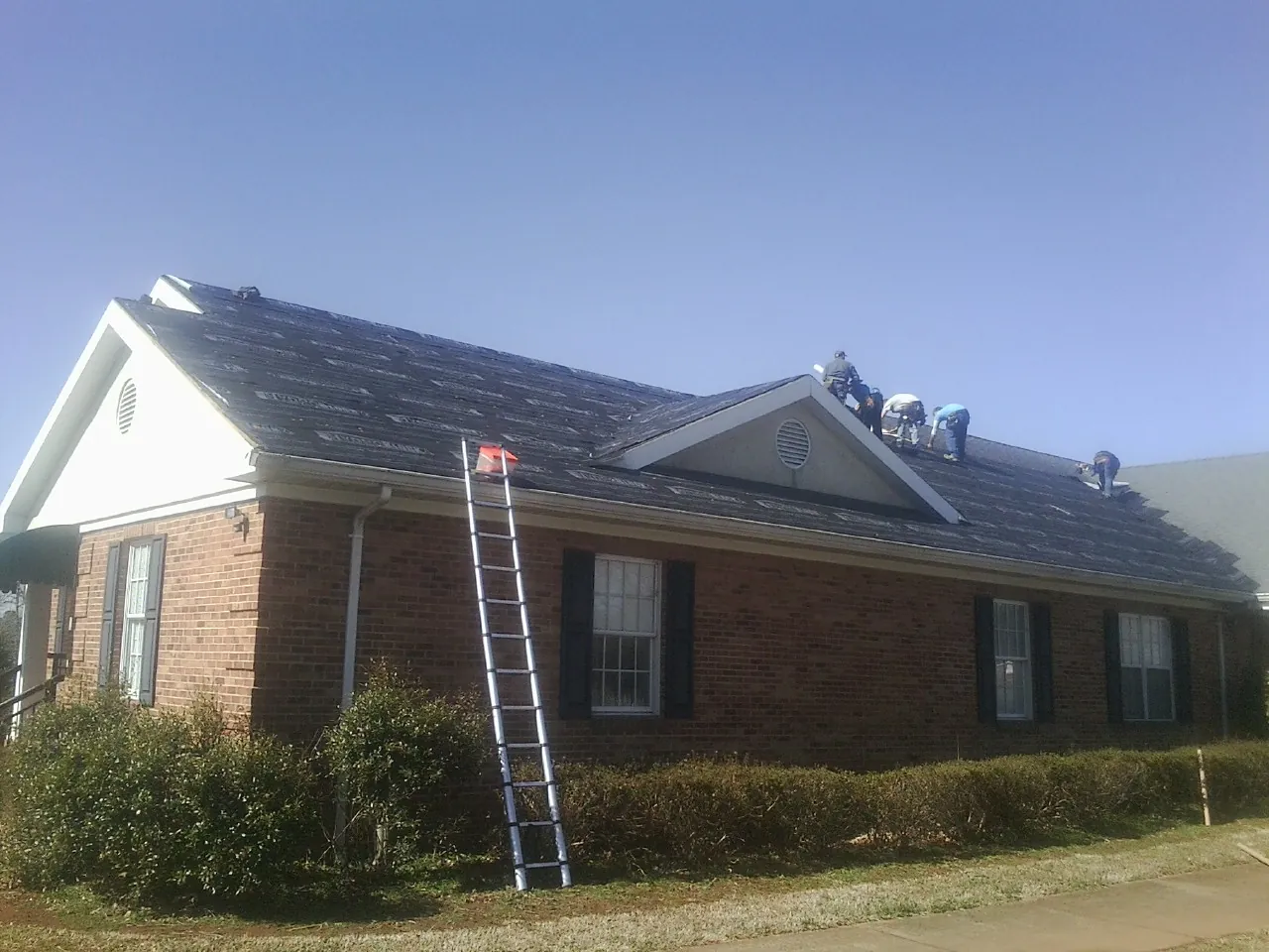 Crew tearing shingles off a brick ranch roof with a ladder set — Emergency Roof Repair work by our crew in Lee Center